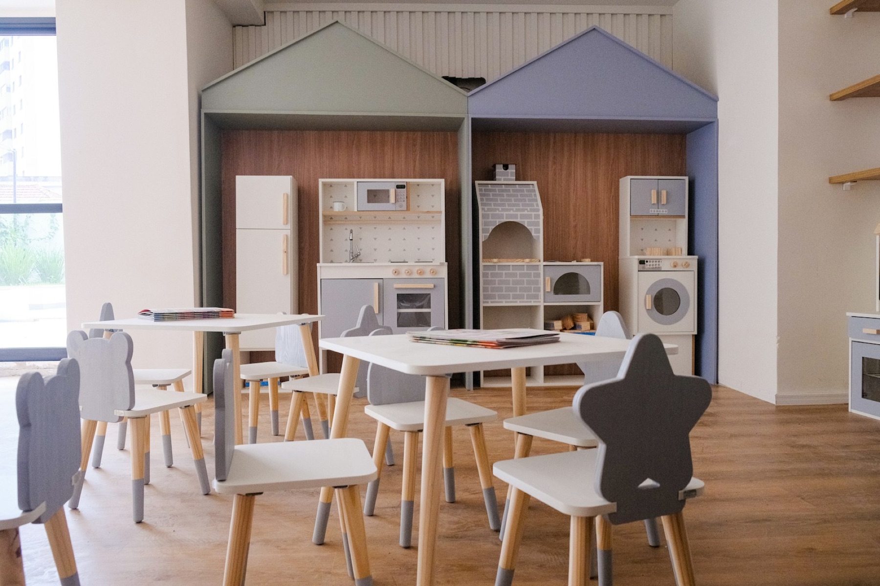 A quiet preschool room with small wooden tables and chairs and a play kitchen